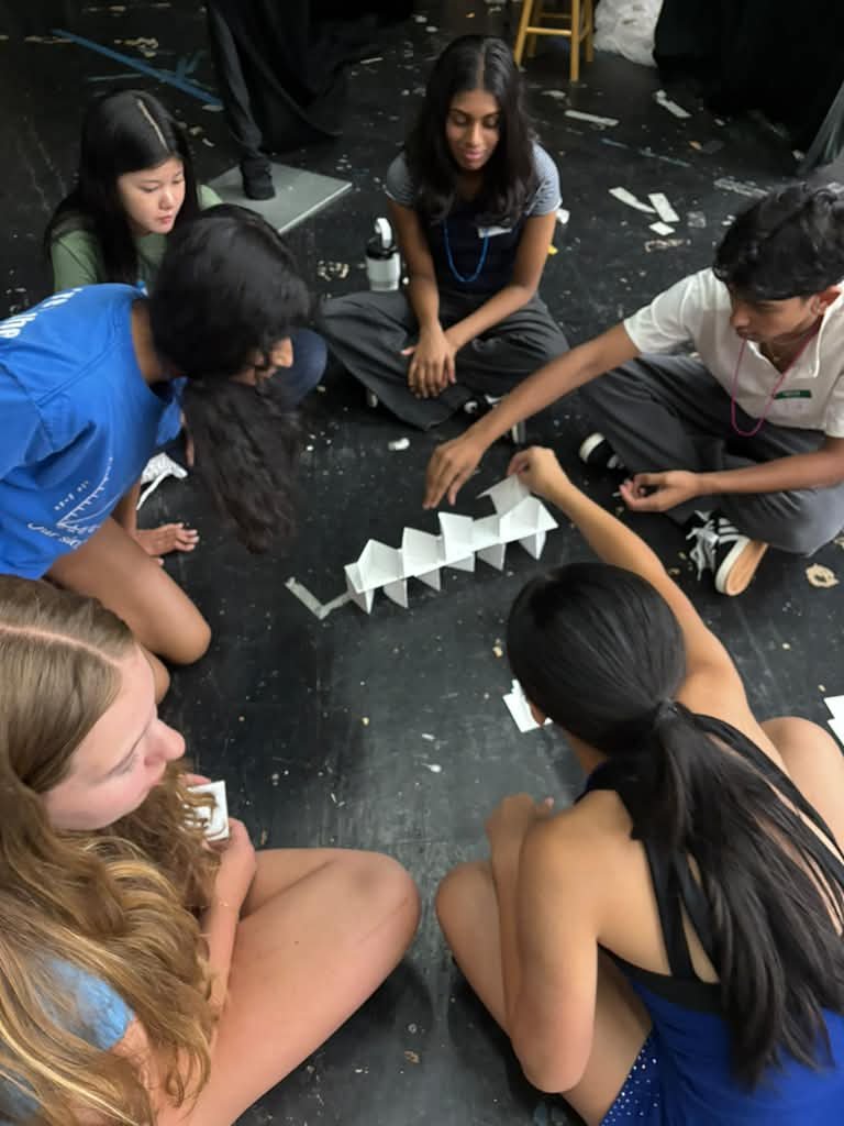 Teens leaning over a zigzag paper-card structure on the stage floor