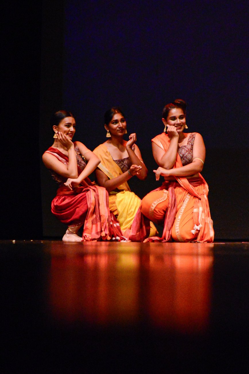 Three Vasantsena dancers posing together in orange and red costumes