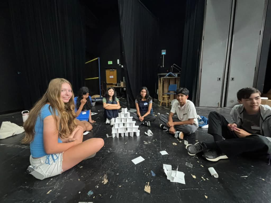 Teens sitting on a dark stage around a small paper-card tower