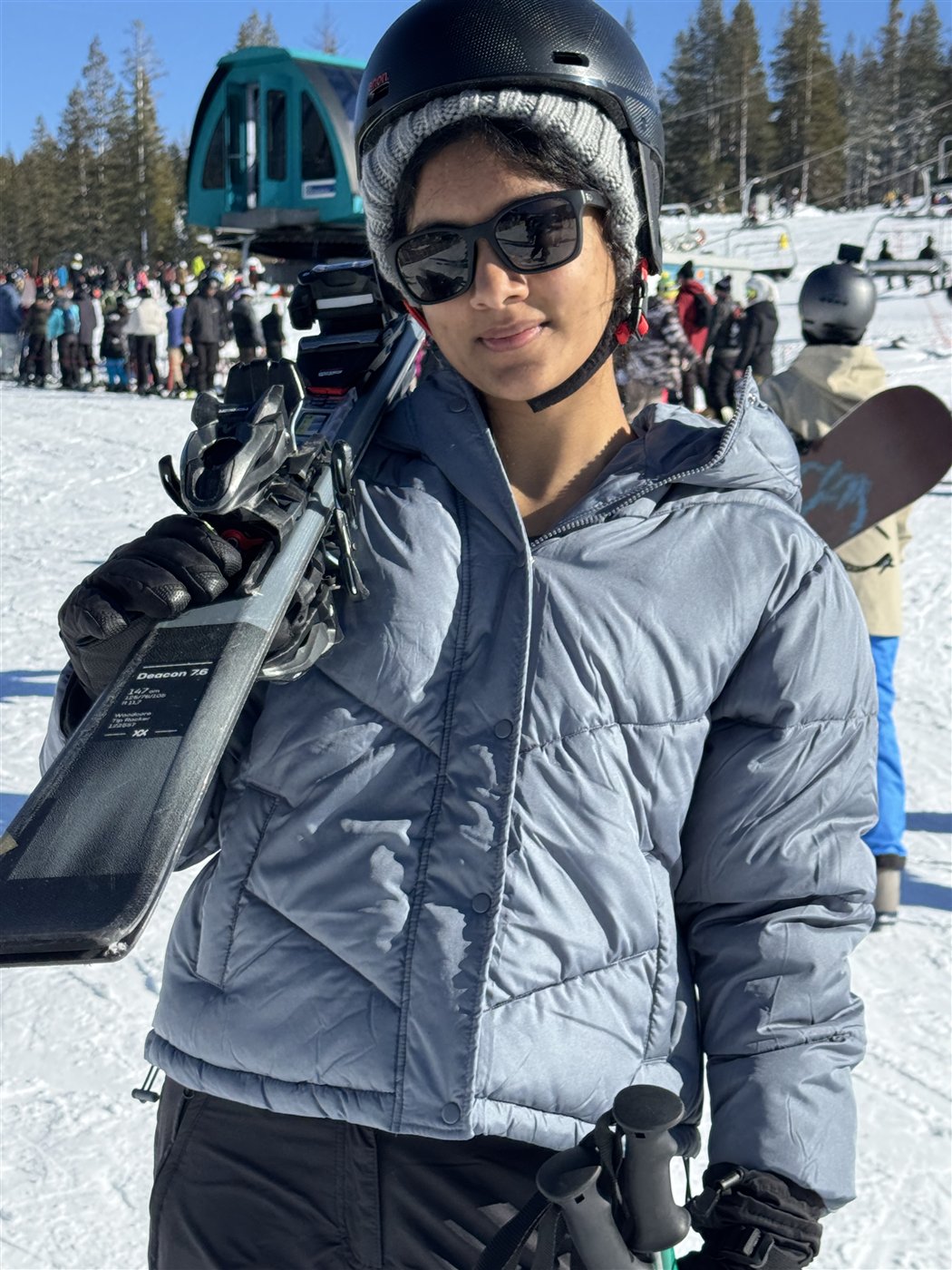Neti in a helmet, sunglasses and puffy jacket with her skis on her shoulder at a ski resort