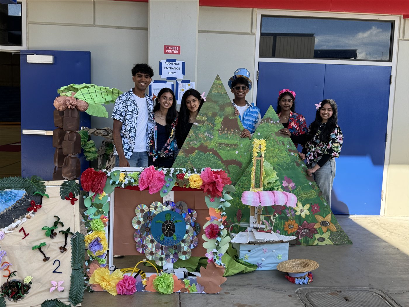 The 2026 DI team standing behind a hand-built tropical set with painted hills, flowers and a CD clock