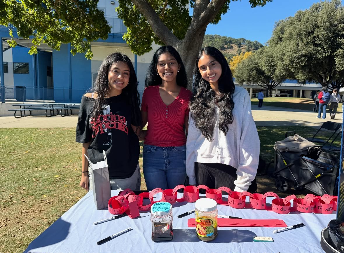 Three TUPE club members smiling at an outdoor awareness booth with a red paper chain